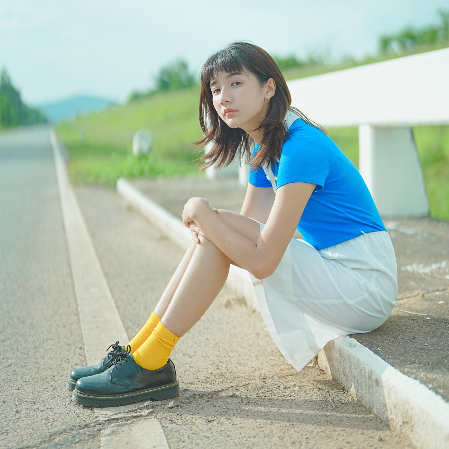 Girl in a bright blue top sitting by the kerb side along an empty road, with her arms over her knees