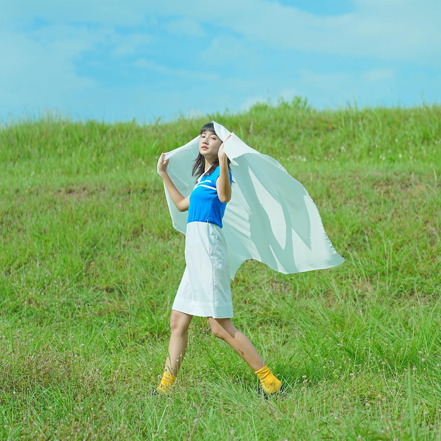 Girl walking in a vast green field, with her eyes closed and holding a white cloth over her head