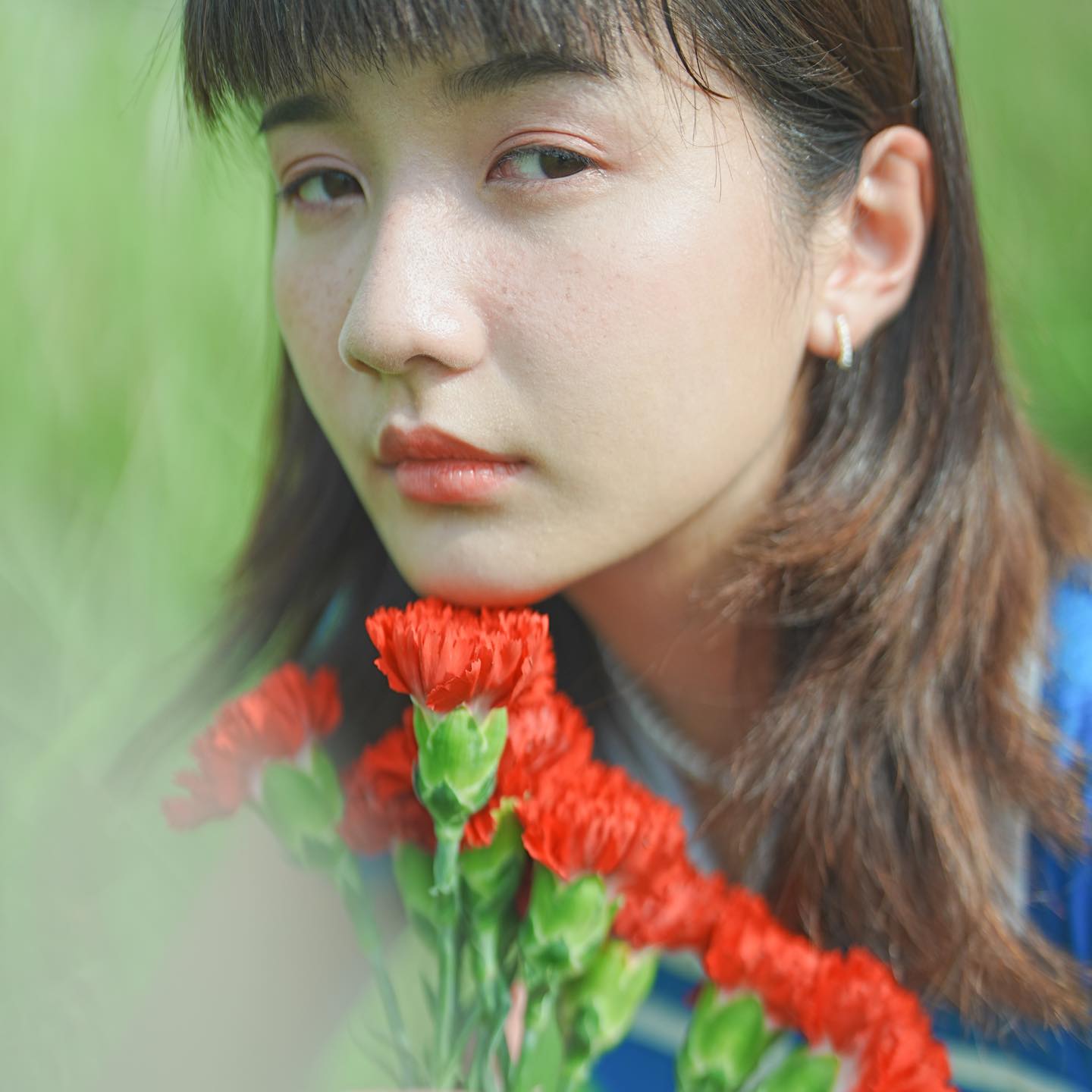 Close-up of girl with bangs and light make-up looking at the camera, with a bunch of red flowers in front of her