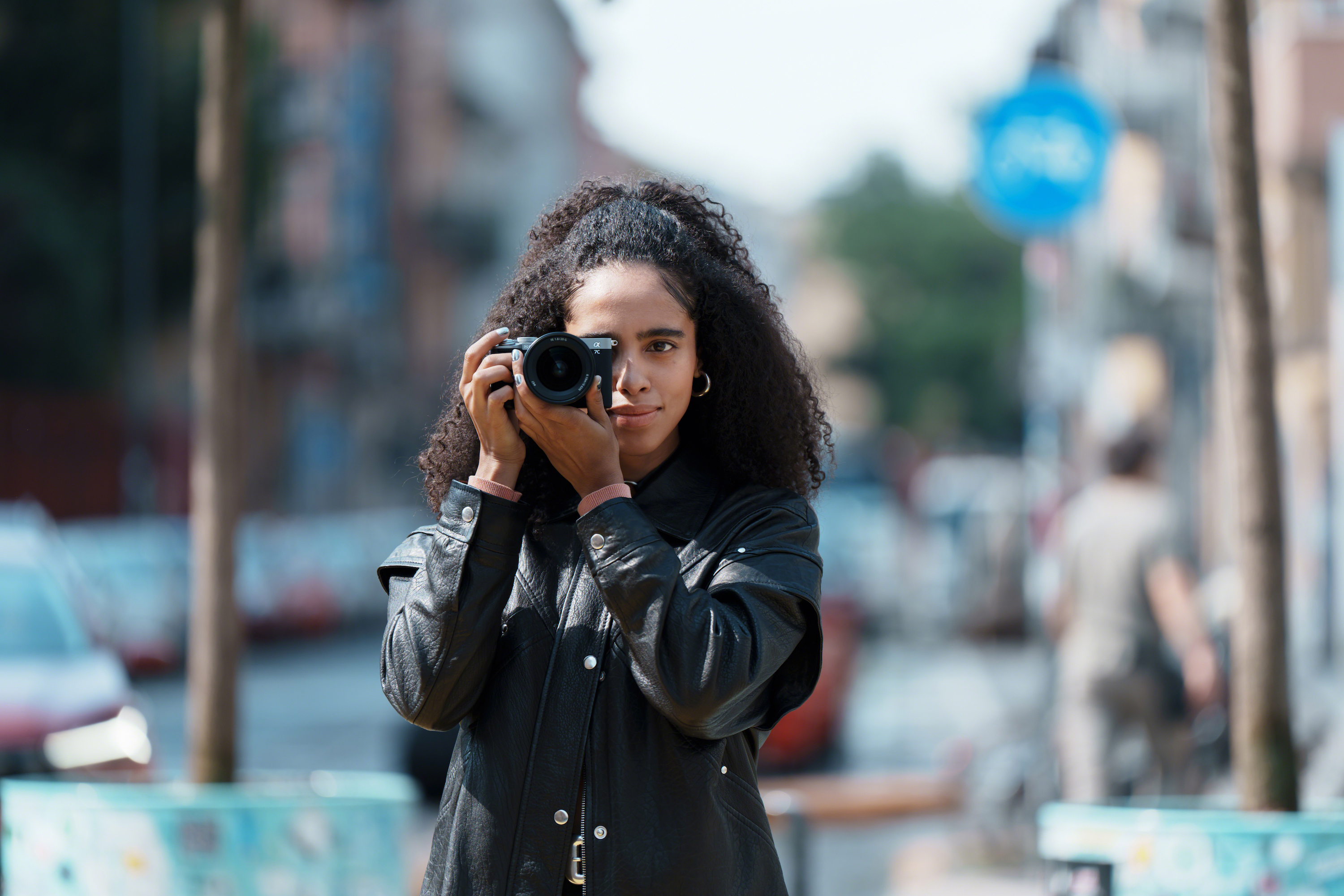 Frontal shot of a girl in a black leather jacket holding up a Sony Alpha 7C to capture a photo
