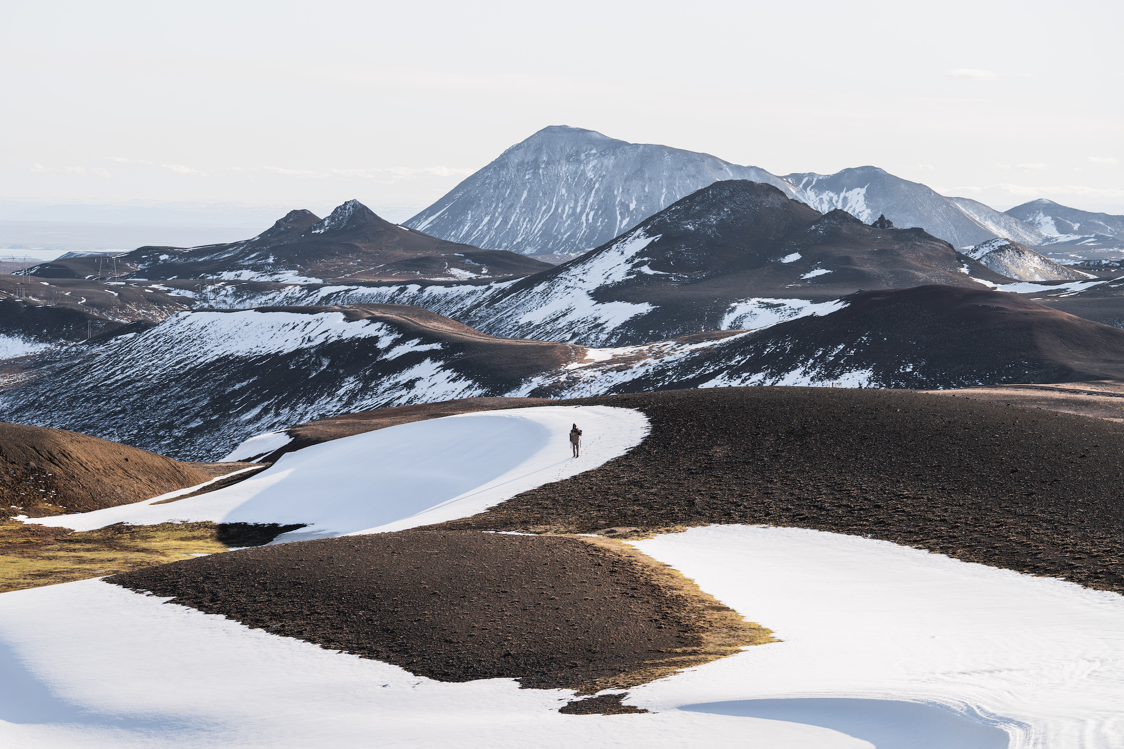 Wide shot of the mountainous landscape with portions covered by snow and a lone traveller trekking