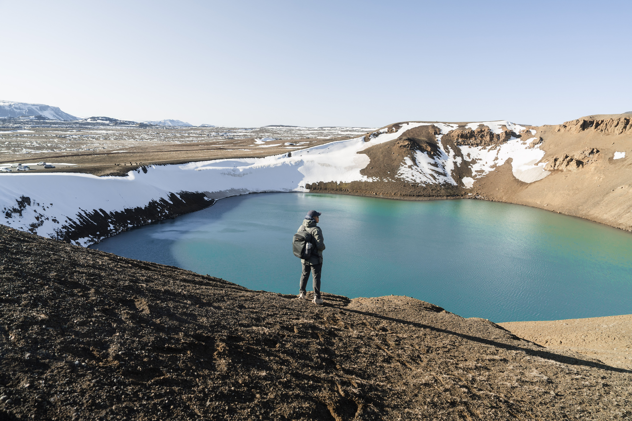 Man in a backpack and coat standing in front of a small alpine lake