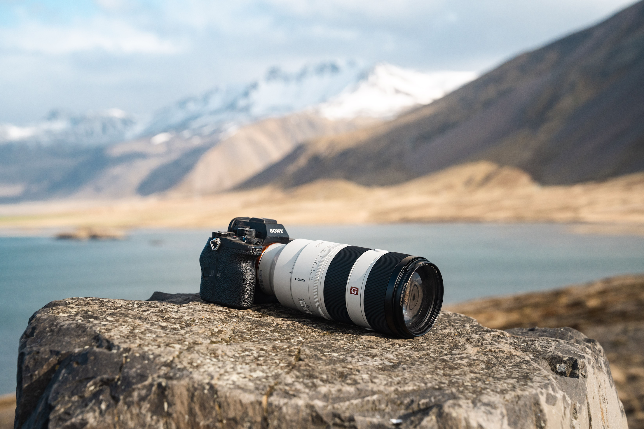 Sony Alpha 7 IV with a zoom lens placed on a rock with a lake and mountains in the background