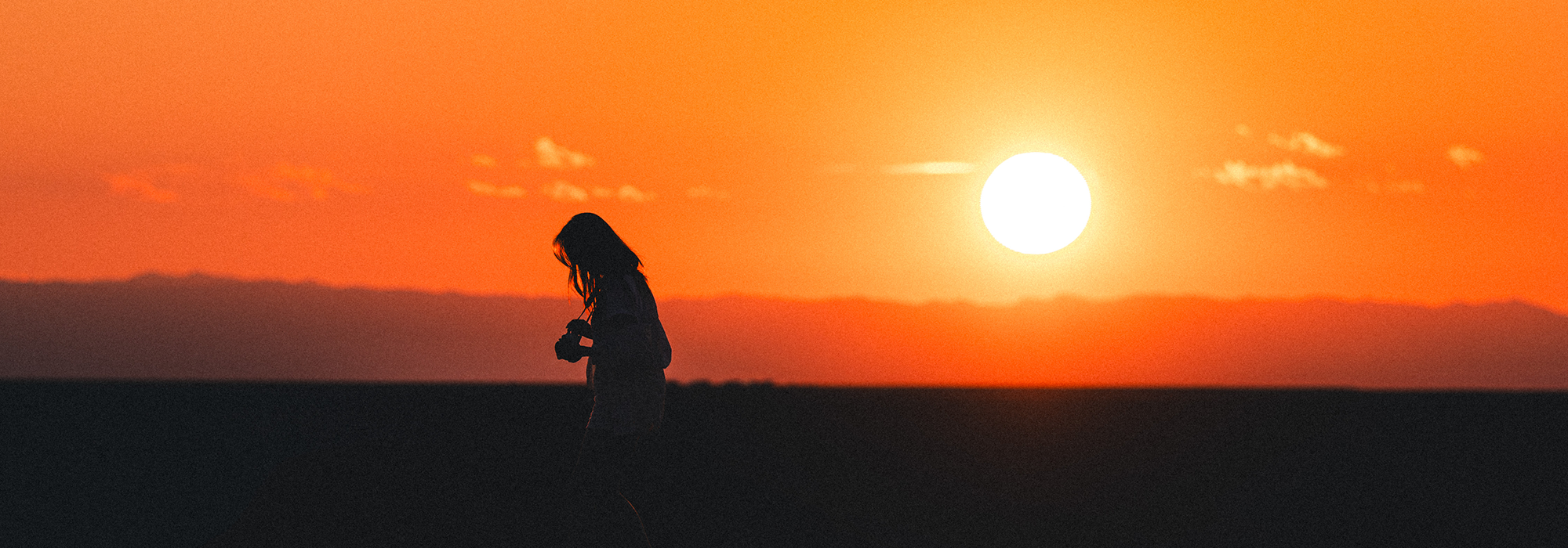 Silhouette of female holding a camera and walking with the setting sun in the background