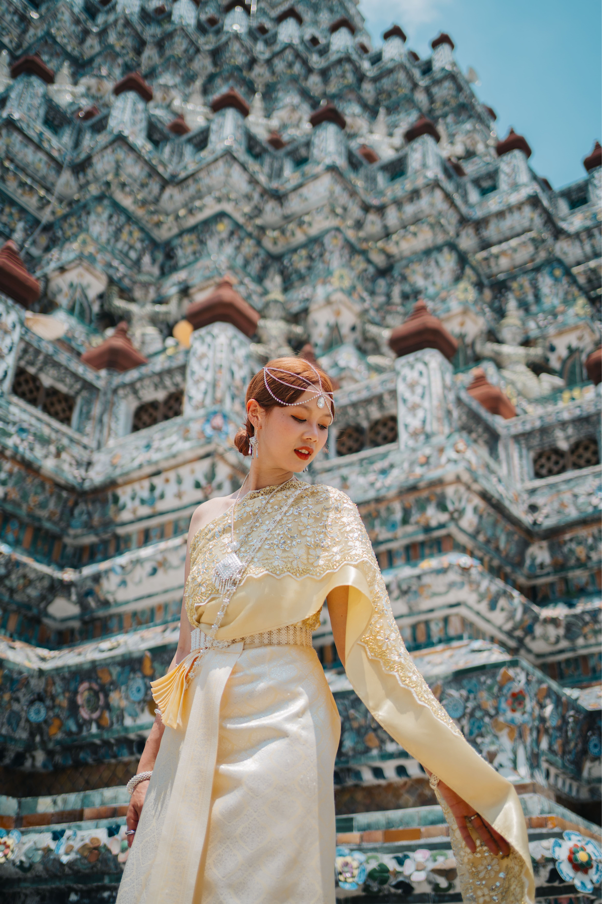 Woman in a traditional Thai outfit looking downwards with her shawl resting on her arm, with the Wat Arun temple in the background