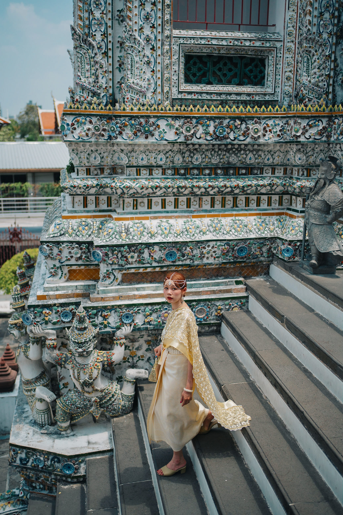 Woman in a traditional Thai outfit walking down the stairs and looking sideways, with the Wat Arun Temple in the background